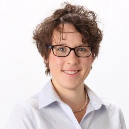 A woman with curly brown hair, wearing glasses, a necklace, and a white collared shirt, smiling against a white background