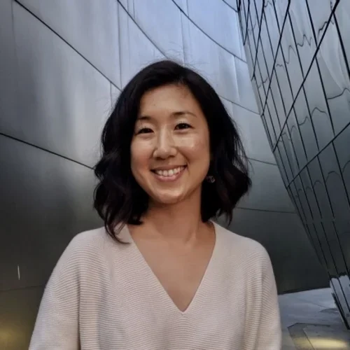 A woman with wavy black hair, wearing a biege sweater, in a room of reflective tiles