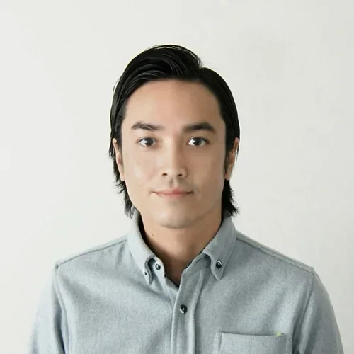A man with medium length black hair, wearing a grey shirt, smiling in front of a beige background