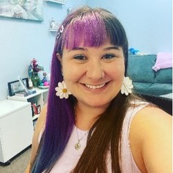 A woman with dyed purple hair, wearing flower earrings and a pink top, smiling in an office