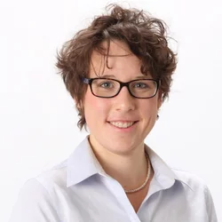 A woman with curly brown hair, wearing glasses, a necklace, and a white collared shirt, smiling against a white background