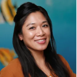 A woman with long black hair, wearing a burnt orange sweater, smiling in front of an aquatic background