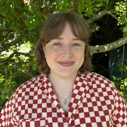 A woman with shoulder length brown hair, wearing a red and white checkered shirt, smiling in front of a forested background