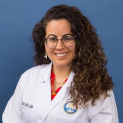 A woman with long curly hair and glasses, wearing a white doctors coat